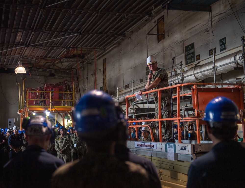 A military officer in camouflage uniform and white hard hat stands elevated on an orange JLG scissor lift, addressing a large crowd of workers and personnel wearing blue hard hats inside what appears to be a large industrial facility or ship hangar bay. The speaker is holding a microphone and appears to be delivering a briefing or address. The setting shows extensive industrial infrastructure including pipes, scaffolding, and overhead lighting.