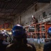 A military officer in camouflage uniform and white hard hat stands elevated on an orange JLG scissor lift, addressing a large crowd of workers and personnel wearing blue hard hats inside what appears to be a large industrial facility or ship hangar bay. The speaker is holding a microphone and appears to be delivering a briefing or address. The setting shows extensive industrial infrastructure including pipes, scaffolding, and overhead lighting.