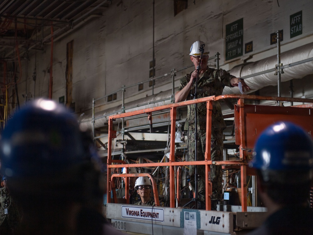 A military personnel member in camouflage uniform and white hard hat stands elevated on a Virginia Equipment JLG scissor lift, appearing to address a group of workers wearing blue hard hats. The setting appears to be inside a large industrial or shipbuilding facility, with visible piping, ductwork, and AFFF sprinkler system signage on the walls. A second person in a white hard hat is visible on the lift platform below the speaker.
