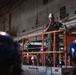 A military personnel member in camouflage uniform and white hard hat stands elevated on a Virginia Equipment JLG scissor lift, appearing to address a group of workers wearing blue hard hats. The setting appears to be inside a large industrial or shipbuilding facility, with visible piping, ductwork, and AFFF sprinkler system signage on the walls. A second person in a white hard hat is visible on the lift platform below the speaker.