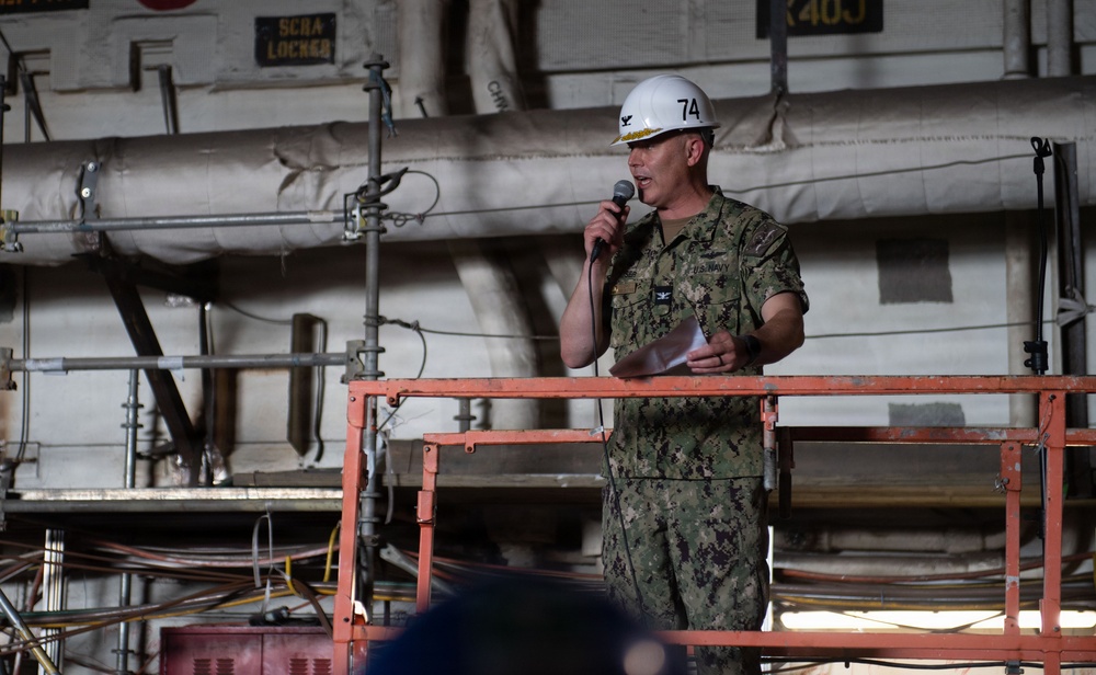A U.S. Navy officer in camouflage uniform (NWU Type III) stands on an elevated red metal platform/scaffold, speaking into a microphone while holding papers. The officer is wearing a white hard hat with the number '74' printed on it and displays a gold eagle insignia indicating the rank of Captain (O-6). The setting appears to be the interior of a naval vessel, likely a hangar bay or industrial shipboard space undergoing maintenance or refit.