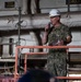A U.S. Navy officer in camouflage uniform (NWU Type III) stands on an elevated red metal platform/scaffold, speaking into a microphone while holding papers. The officer is wearing a white hard hat with the number '74' printed on it and displays a gold eagle insignia indicating the rank of Captain (O-6). The setting appears to be the interior of a naval vessel, likely a hangar bay or industrial shipboard space undergoing maintenance or refit.