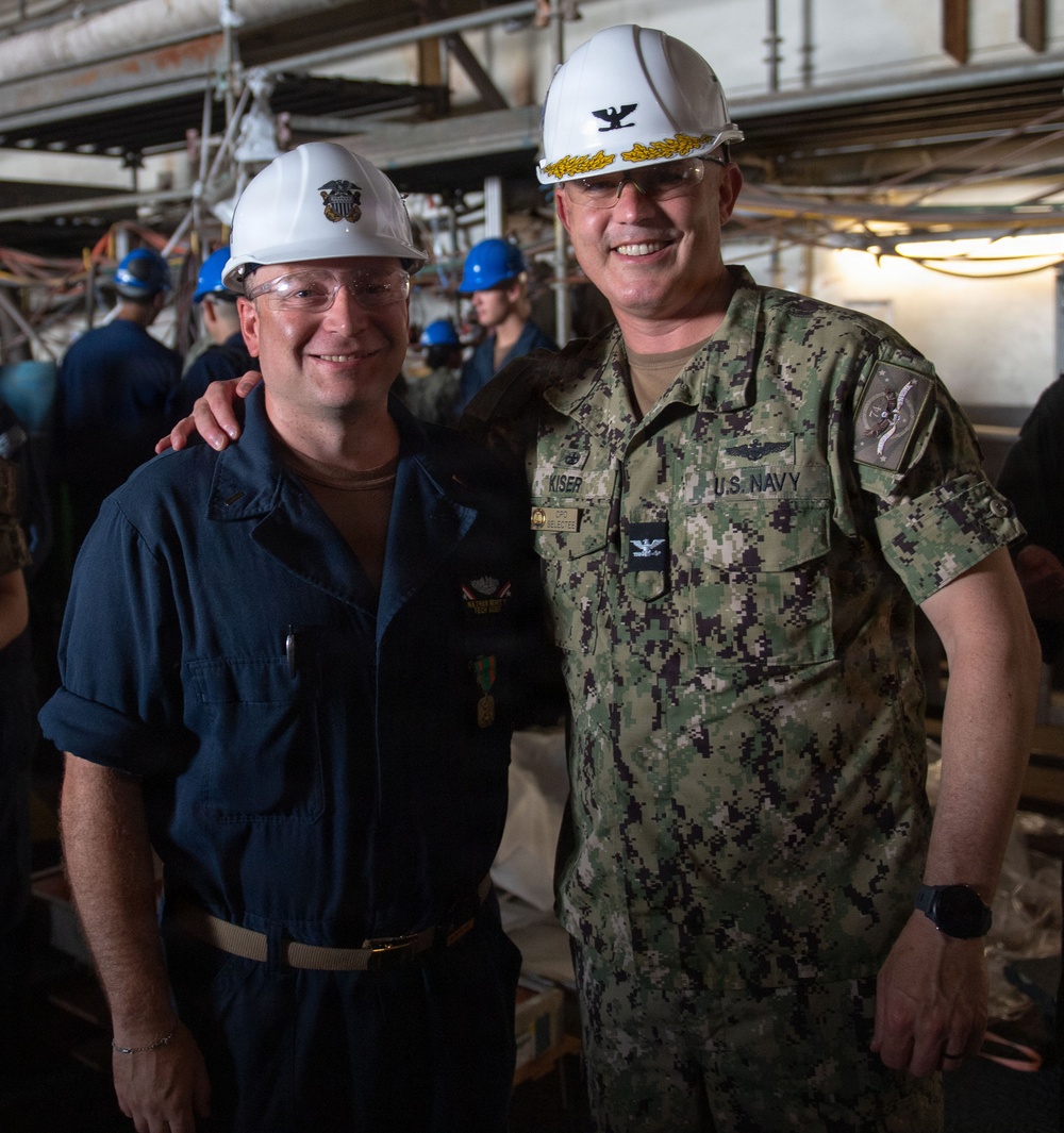 Two U.S. Navy personnel pose together for a photo in what appears to be a ship's interior undergoing maintenance or repair work. The individual on the right is identified by his name tape as 'KISER' and wears a Captain's rank insignia (eagle) on his white hard hat along with gold oak leaf scrambled eggs, indicating senior officer status. Both wear white hard hats and are surrounded by workers in blue hard hats in a industrial shipboard environment.