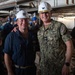 Two U.S. Navy personnel pose together for a photo in what appears to be a ship's interior undergoing maintenance or repair work. The individual on the right is identified by his name tape as 'KISER' and wears a Captain's rank insignia (eagle) on his white hard hat along with gold oak leaf scrambled eggs, indicating senior officer status. Both wear white hard hats and are surrounded by workers in blue hard hats in a industrial shipboard environment.
