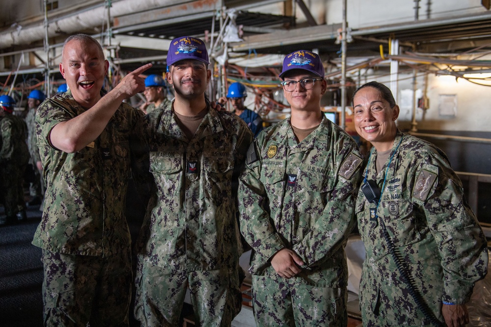 Four U.S. Navy personnel in NWU Type III (digital camouflage) uniforms pose for a group photo inside what appears to be a naval vessel's interior workspace. Two of the sailors are wearing purple baseball caps with an embroidered ship or command insignia. The leftmost individual is gesturing playfully toward one of the cap-wearers, suggesting a celebratory or recognition moment.