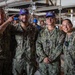 Four U.S. Navy personnel in NWU Type III (digital camouflage) uniforms pose for a group photo inside what appears to be a naval vessel's interior workspace. Two of the sailors are wearing purple baseball caps with an embroidered ship or command insignia. The leftmost individual is gesturing playfully toward one of the cap-wearers, suggesting a celebratory or recognition moment.