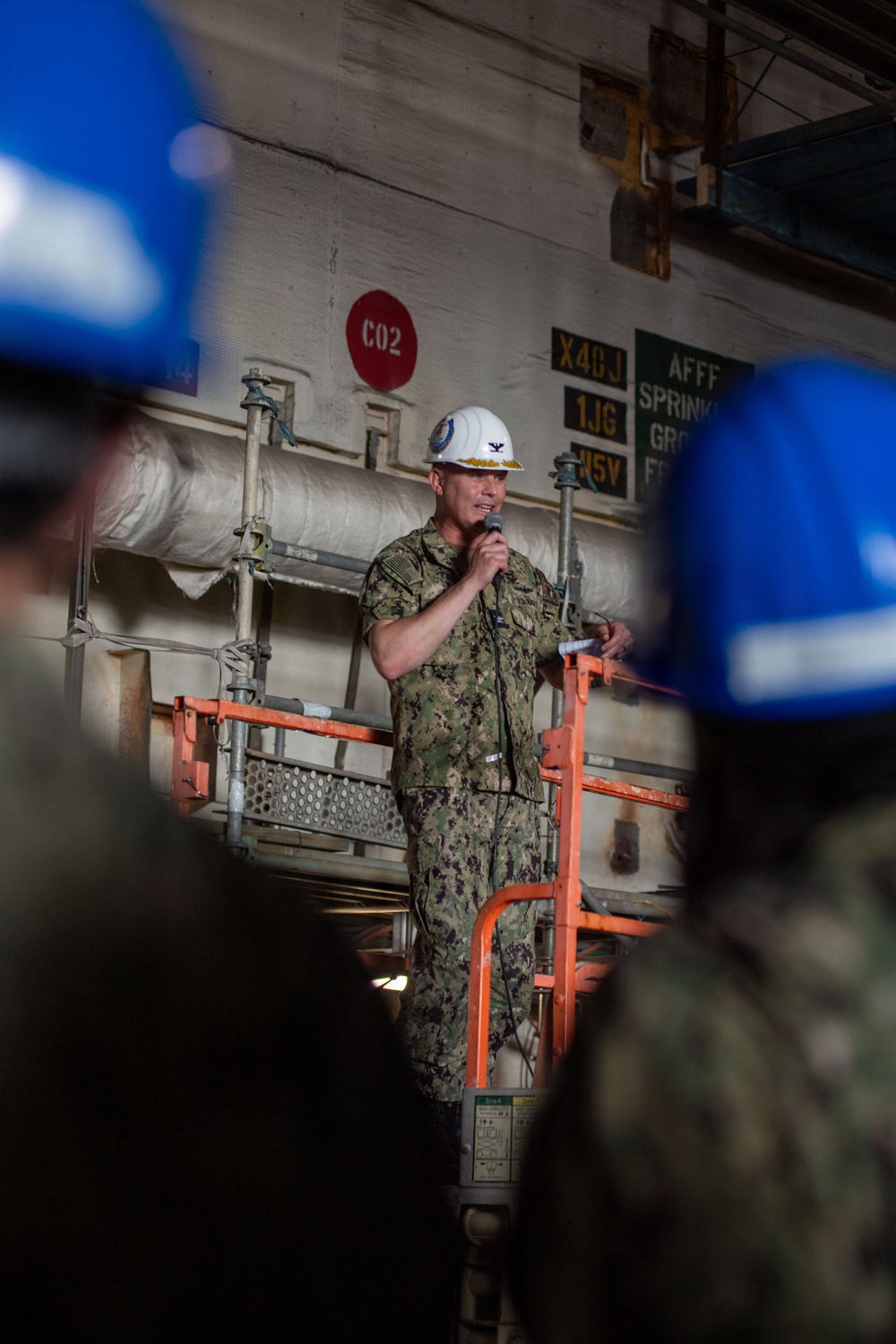A U.S. Navy officer in camouflage NWU (Navy Working Uniform) and a white hard hat is speaking into a microphone while standing on or near an orange scissor lift platform inside what appears to be a naval vessel or shipyard facility. The officer appears to be addressing a group of personnel, several of whom are wearing blue hard hats visible in the foreground. Signage on the bulkhead behind includes CO2 markings and AFFF sprinkler system labels, consistent with a naval ship interior.