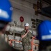 A U.S. Navy officer in camouflage NWU (Navy Working Uniform) and a white hard hat is speaking into a microphone while standing on or near an orange scissor lift platform inside what appears to be a naval vessel or shipyard facility. The officer appears to be addressing a group of personnel, several of whom are wearing blue hard hats visible in the foreground. Signage on the bulkhead behind includes CO2 markings and AFFF sprinkler system labels, consistent with a naval ship interior.
