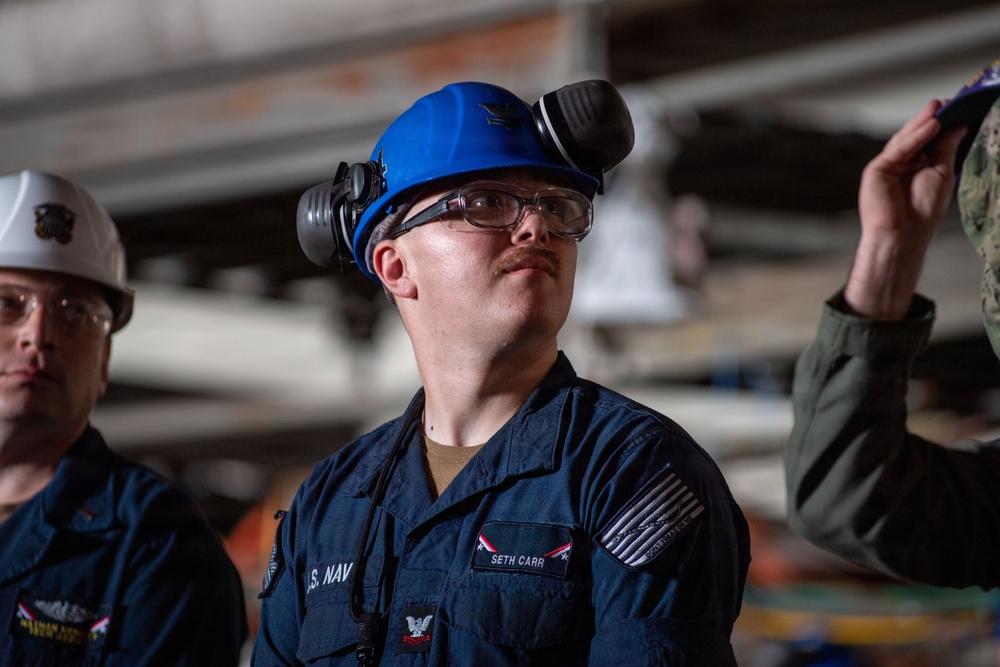 A U.S. Navy sailor named Seth Carr, wearing a blue hard hat with ear protection and safety glasses, is looking upward in what appears to be an industrial or shipboard environment. He is in Navy working uniform (NWU) with a name tape and rank insignia visible. At least two other personnel are partially visible in the background.