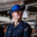 A U.S. Navy sailor named Seth Carr, wearing a blue hard hat with ear protection and safety glasses, is looking upward in what appears to be an industrial or shipboard environment. He is in Navy working uniform (NWU) with a name tape and rank insignia visible. At least two other personnel are partially visible in the background.