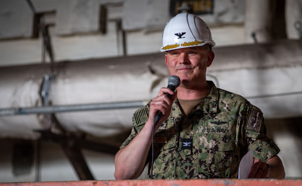 A U.S. Navy officer wearing digital camouflage NWU (Navy Working Uniform) and a white hard hat with gold oak leaf scrambled eggs and an eagle insignia is speaking into a handheld microphone. The setting appears to be an industrial or shipboard environment, likely a hangar bay or dry dock facility. The officer holds papers in one hand, suggesting a formal address or ceremony.