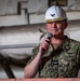 A U.S. Navy officer wearing digital camouflage NWU (Navy Working Uniform) and a white hard hat with gold oak leaf scrambled eggs and an eagle insignia is speaking into a handheld microphone. The setting appears to be an industrial or shipboard environment, likely a hangar bay or dry dock facility. The officer holds papers in one hand, suggesting a formal address or ceremony.