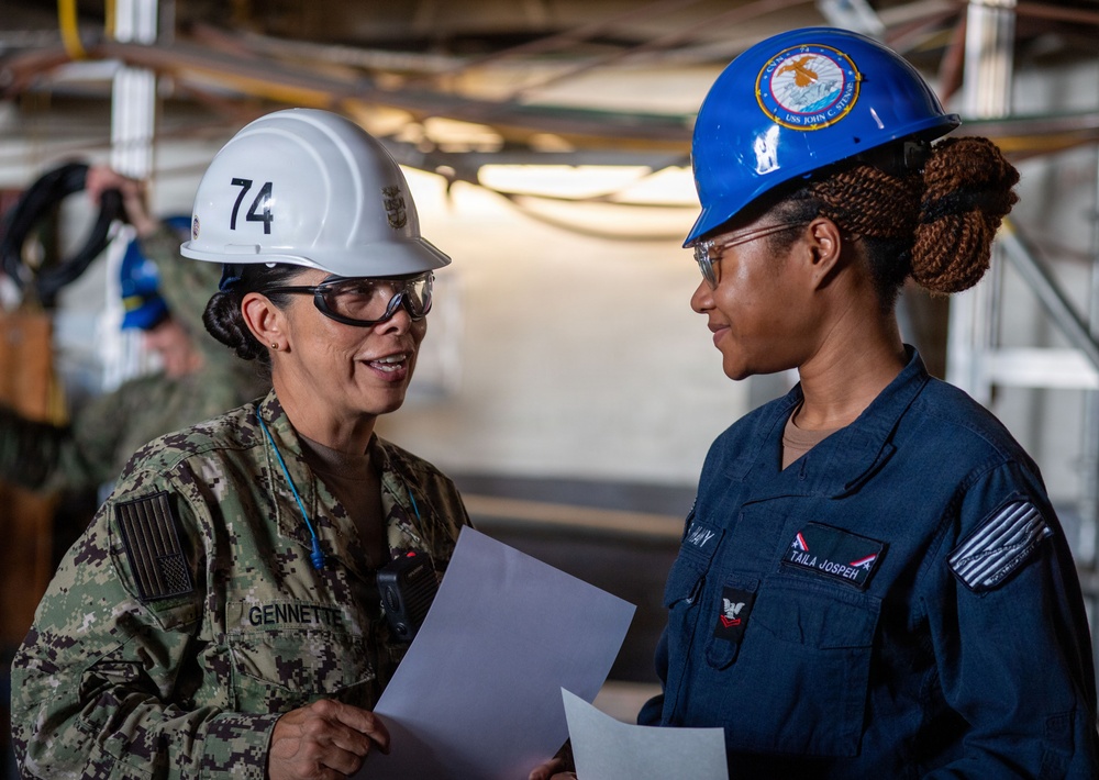 Two female U.S. Navy personnel are engaged in a work-related discussion in an industrial or shipboard environment. The individual on the left wears camouflage NWU (Navy Working Uniform) with a white hard hat numbered '74' and the name tag 'GENNETTE', while the individual on the right wears a blue coverall uniform with the name tag 'TAILA JOSPEH' and a blue hard hat bearing a USS John C. Stennis ship insignia. Both are holding documents and appear to be reviewing or discussing work-related materials.
