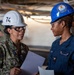 Two female U.S. Navy personnel are engaged in a work-related discussion in an industrial or shipboard environment. The individual on the left wears camouflage NWU (Navy Working Uniform) with a white hard hat numbered '74' and the name tag 'GENNETTE', while the individual on the right wears a blue coverall uniform with the name tag 'TAILA JOSPEH' and a blue hard hat bearing a USS John C. Stennis ship insignia. Both are holding documents and appear to be reviewing or discussing work-related materials.
