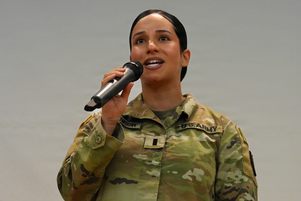 A female U.S. Army soldier in Operational Camouflage Pattern (OCP) uniform is speaking or performing into a handheld microphone. She appears to be addressing an audience or singing at a military event. Her uniform displays a 'U.S. ARMY' tape, a name tape partially visible, and what appears to be a First Lieutenant rank insignia.