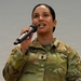 A female U.S. Army soldier in Operational Camouflage Pattern (OCP) uniform is speaking or performing into a handheld microphone. She appears to be addressing an audience or singing at a military event. Her uniform displays a 'U.S. ARMY' tape, a name tape partially visible, and what appears to be a First Lieutenant rank insignia.