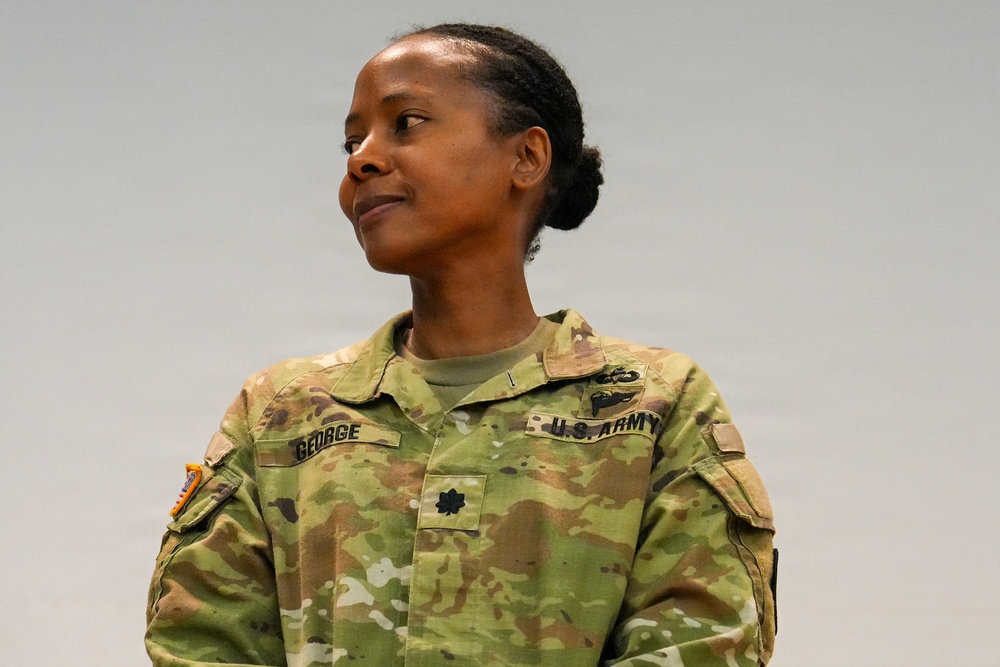 A U.S. Army soldier wearing Operational Camouflage Pattern (OCP) uniform is photographed against a neutral gray background. The name tape reads 'GEORGE' and the rank insignia on the chest indicates the rank of Major (O-4), identified by the black oak leaf cluster. A medical corps branch insignia is visible on the right chest area.