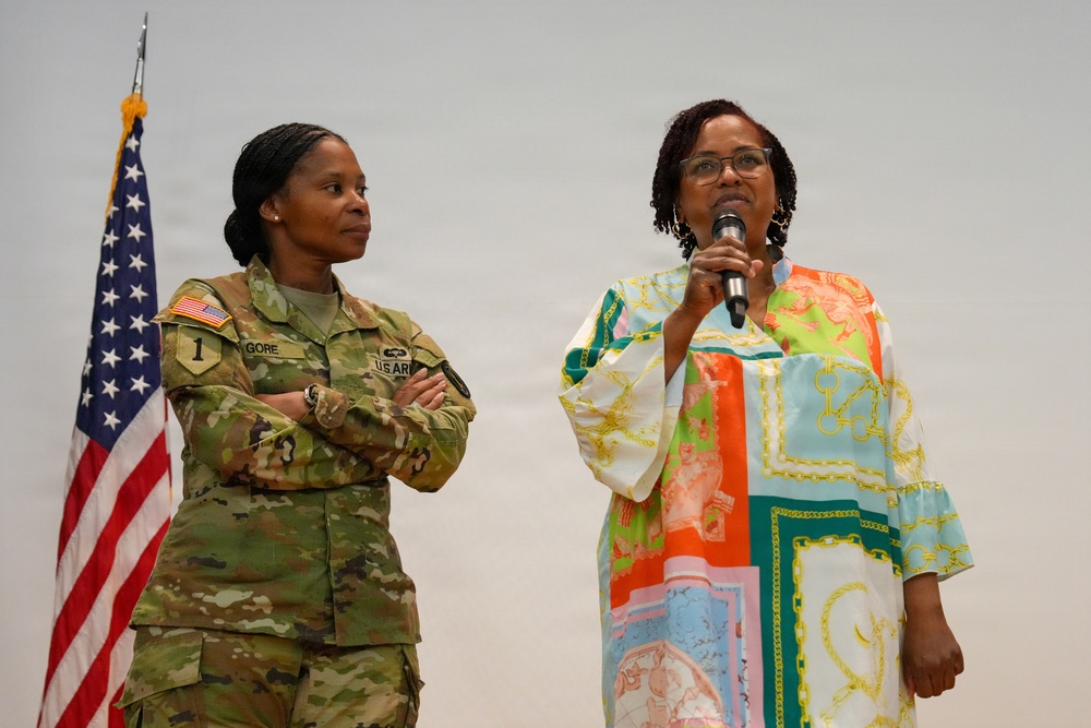 Two women are standing together at what appears to be a military event or ceremony. One woman is wearing a U.S. Army camouflage uniform with the name tape 'GORE' and a '1' patch, while the other is speaking into a microphone wearing a colorful patterned dress. An American flag is visible in the background to the left.