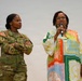 Two women are standing together at what appears to be a military event or ceremony. One woman is wearing a U.S. Army camouflage uniform with the name tape 'GORE' and a '1' patch, while the other is speaking into a microphone wearing a colorful patterned dress. An American flag is visible in the background to the left.