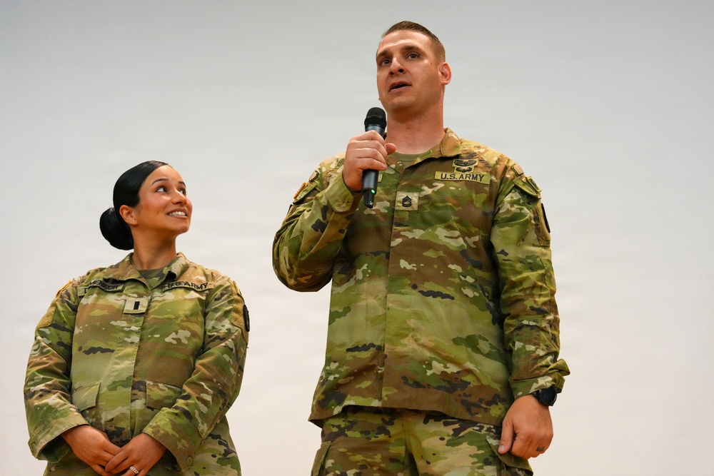 Two U.S. Army soldiers in Operational Camouflage Pattern (OCP) uniforms are standing together, with the male soldier speaking into a microphone. The female soldier stands to his left, smiling and looking up at him. The setting appears to be an indoor military event or ceremony.