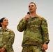 Two U.S. Army soldiers in Operational Camouflage Pattern (OCP) uniforms are standing together, with the male soldier speaking into a microphone. The female soldier stands to his left, smiling and looking up at him. The setting appears to be an indoor military event or ceremony.