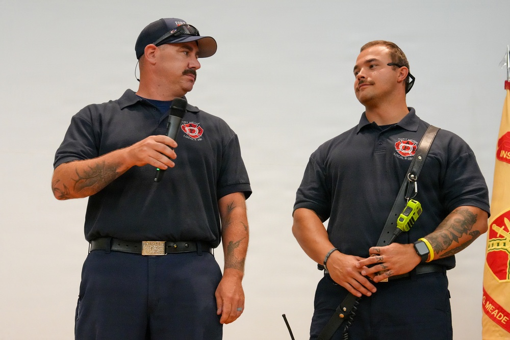 Two firefighters in matching navy blue uniform polo shirts with a Local fire department emblem are standing together, likely at a formal presentation or ceremony. The man on the left is speaking into a handheld microphone while the younger man on the right listens attentively. A military or organizational flag is partially visible in the background on the right.
