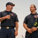 Two firefighters in matching navy blue uniform polo shirts with a Local fire department emblem are standing together, likely at a formal presentation or ceremony. The man on the left is speaking into a handheld microphone while the younger man on the right listens attentively. A military or organizational flag is partially visible in the background on the right.