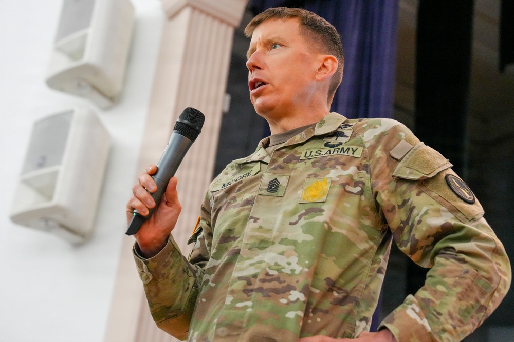 A U.S. Army soldier in OCP (Operational Camouflage Pattern) uniform is speaking into a handheld microphone, appearing to address an audience. The name tape on the uniform reads 'MOORE' and rank insignia indicates a senior non-commissioned officer. The setting appears to be an indoor venue or auditorium.