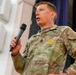 A U.S. Army soldier in OCP (Operational Camouflage Pattern) uniform is speaking into a handheld microphone, appearing to address an audience. The name tape on the uniform reads 'MOORE' and rank insignia indicates a senior non-commissioned officer. The setting appears to be an indoor venue or auditorium.
