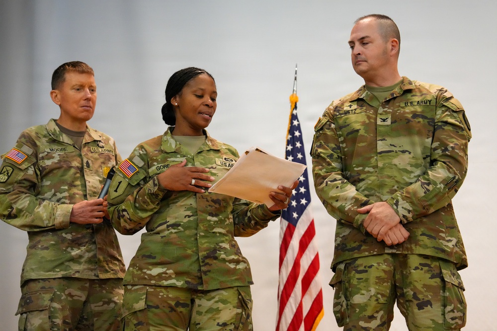 Three U.S. Army soldiers in MultiCam (OCP) uniforms stand before an American flag during what appears to be a formal military ceremony. The central soldier, identified by name tape as 'GORE,' is reading from a document while the soldier on the left (MOORE) holds a pen and the soldier on the right (METZ) stands at ease. The presence of a document, flag, and formal posture suggests this is a promotion, award, or oath-of-office ceremony.