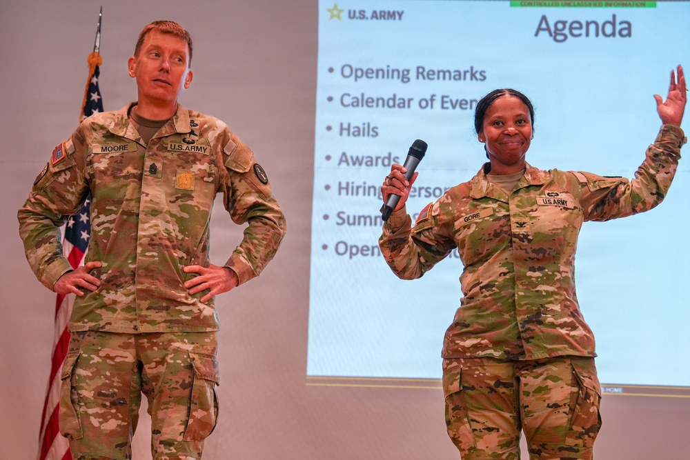 Two U.S. Army soldiers in OCP (Operational Camouflage Pattern) uniforms are presenting at a formal military briefing or meeting. The soldier on the right, identified by nametape as 'GORE,' is holding a microphone and gesturing with one hand raised, while the soldier on the left, identified as 'MOORE,' stands with hands on hips. A projected presentation slide displaying a U.S. Army agenda is visible in the background.