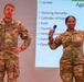 Two U.S. Army soldiers in OCP (Operational Camouflage Pattern) uniforms are presenting at a formal military briefing or meeting. The soldier on the right, identified by nametape as 'GORE,' is holding a microphone and gesturing with one hand raised, while the soldier on the left, identified as 'MOORE,' stands with hands on hips. A projected presentation slide displaying a U.S. Army agenda is visible in the background.