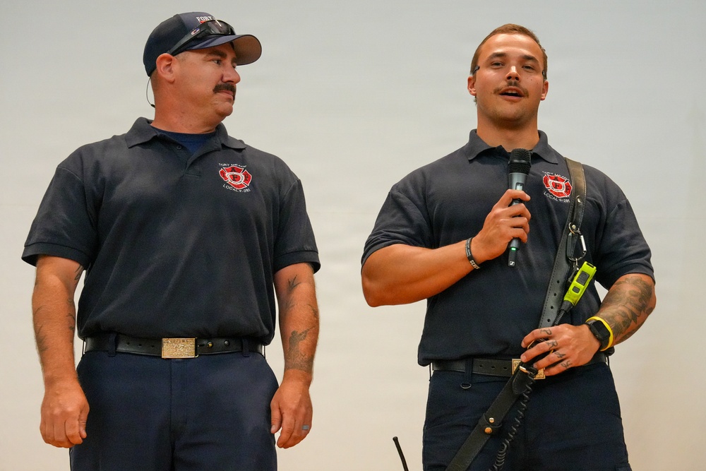 Two men wearing matching navy blue polo shirts with a fire department logo (Fort McCoy Local 281) are standing together, likely at a presentation or public event. The man on the right is speaking into a handheld microphone while holding audio equipment with a strap. Both men have visible tattoos and are dressed in uniform attire consistent with firefighters or fire union representatives.