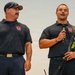 Two men wearing matching navy blue polo shirts with a fire department logo (Fort McCoy Local 281) are standing together, likely at a presentation or public event. The man on the right is speaking into a handheld microphone while holding audio equipment with a strap. Both men have visible tattoos and are dressed in uniform attire consistent with firefighters or fire union representatives.