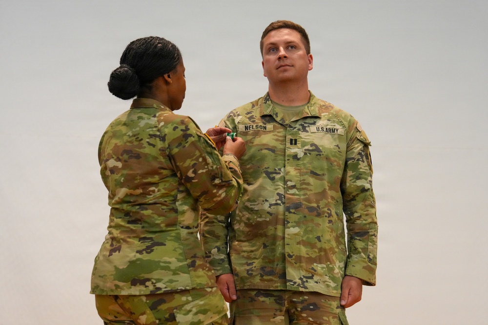 A U.S. Army ceremony is taking place in which one soldier is pinning a medal or insignia onto another soldier's uniform. The recipient, identified by the name tape 'NELSON' and wearing captain's rank insignia, stands at attention while facing forward. Both soldiers are wearing the Army Combat Uniform (ACU) in OCP (Operational Camouflage Pattern).