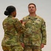 A U.S. Army ceremony is taking place in which one soldier is pinning a medal or insignia onto another soldier's uniform. The recipient, identified by the name tape 'NELSON' and wearing captain's rank insignia, stands at attention while facing forward. Both soldiers are wearing the Army Combat Uniform (ACU) in OCP (Operational Camouflage Pattern).