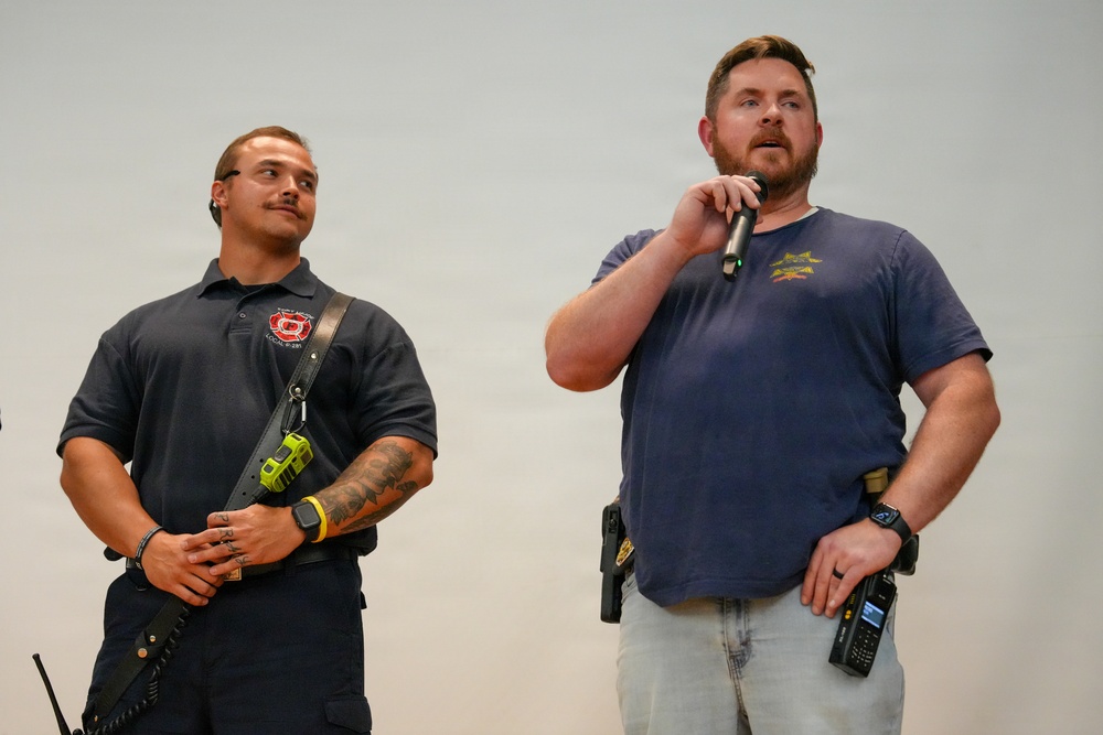 Two men are standing in front of a plain light-colored background, appearing to be at a public event or presentation. The man on the right is speaking into a handheld microphone while the man on the left listens attentively. Both men appear to be in public safety roles based on their attire and equipment.