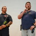 Two men are standing in front of a plain light-colored background, appearing to be at a public event or presentation. The man on the right is speaking into a handheld microphone while the man on the left listens attentively. Both men appear to be in public safety roles based on their attire and equipment.