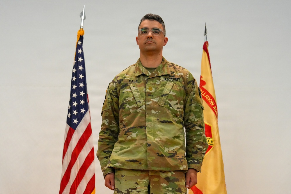 A U.S. Army soldier named Cale stands at attention in an official military portrait setting. He is wearing the Operational Camouflage Pattern (OCP) uniform with a rank insignia visible on his chest. Two flags — the U.S. national flag and what appears to be an Installation Management Command flag — are displayed behind him.