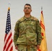 A U.S. Army soldier named Cale stands at attention in an official military portrait setting. He is wearing the Operational Camouflage Pattern (OCP) uniform with a rank insignia visible on his chest. Two flags — the U.S. national flag and what appears to be an Installation Management Command flag — are displayed behind him.