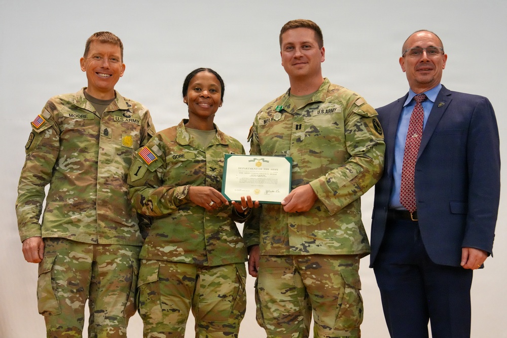 Four individuals pose for a formal photo during a U.S. Army award ceremony. Two soldiers in the center are jointly holding a Department of the Army certificate, flanked by a senior NCO on the left and a civilian official on the right. The setting appears to be an indoor military venue with a plain light-colored background.