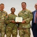 Four individuals pose for a formal photo during a U.S. Army award ceremony. Two soldiers in the center are jointly holding a Department of the Army certificate, flanked by a senior NCO on the left and a civilian official on the right. The setting appears to be an indoor military venue with a plain light-colored background.
