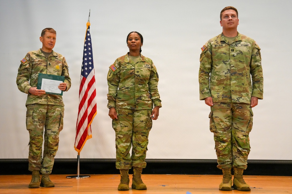 Three U.S. Army soldiers in OCP (Operational Camouflage Pattern) uniforms stand on a stage during what appears to be a military ceremony. An American flag on a stand is positioned between the left and center soldiers. The soldier on the left, identified by nametape as 'MOORE,' holds a green folder or document, suggesting he may be officiating or reading orders.