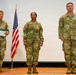 Three U.S. Army soldiers in OCP (Operational Camouflage Pattern) uniforms stand on a stage during what appears to be a military ceremony. An American flag on a stand is positioned between the left and center soldiers. The soldier on the left, identified by nametape as 'MOORE,' holds a green folder or document, suggesting he may be officiating or reading orders.