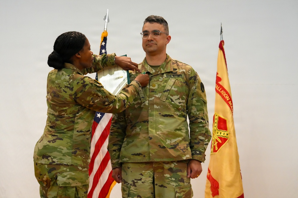 Two U.S. Army soldiers in OCP (Operational Camouflage Pattern) uniforms are participating in a military awards or promotion ceremony. One soldier is pinning a medal or insignia onto the chest of the other soldier, who is standing at attention. An American flag and what appears to be an Installation Management Command guidon are visible in the background.