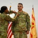 Two U.S. Army soldiers in OCP (Operational Camouflage Pattern) uniforms are participating in a military awards or promotion ceremony. One soldier is pinning a medal or insignia onto the chest of the other soldier, who is standing at attention. An American flag and what appears to be an Installation Management Command guidon are visible in the background.