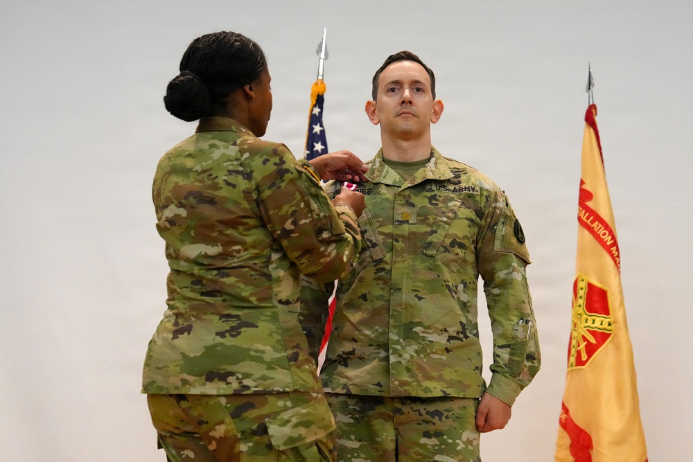 A U.S. Army military ceremony is taking place in which a female soldier is pinning a medal or insignia onto a male soldier's uniform. Both individuals are wearing Army Combat Uniforms (ACU) in OCP (Operational Camouflage Pattern). An American flag and what appears to be an Installation Management Command flag are visible in the background.