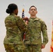 A U.S. Army military ceremony is taking place in which a female soldier is pinning a medal or insignia onto a male soldier's uniform. Both individuals are wearing Army Combat Uniforms (ACU) in OCP (Operational Camouflage Pattern). An American flag and what appears to be an Installation Management Command flag are visible in the background.