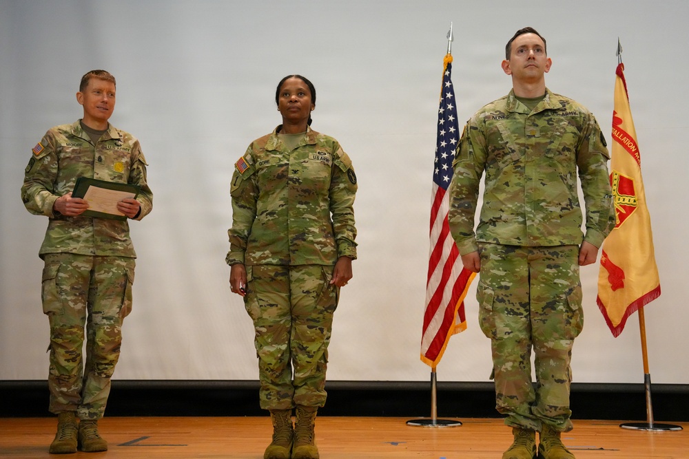 Three U.S. Army soldiers in OCP (Operational Camouflage Pattern) uniforms stand on a stage during what appears to be a military ceremony. Two flags are visible behind them — the American flag and what appears to be a U.S. Army Installation Management Command guidon. The soldier on the left is holding a folder or certificate, suggesting an awards or promotion ceremony.