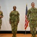 Three U.S. Army soldiers in OCP (Operational Camouflage Pattern) uniforms stand on a stage during what appears to be a military ceremony. Two flags are visible behind them — the American flag and what appears to be a U.S. Army Installation Management Command guidon. The soldier on the left is holding a folder or certificate, suggesting an awards or promotion ceremony.